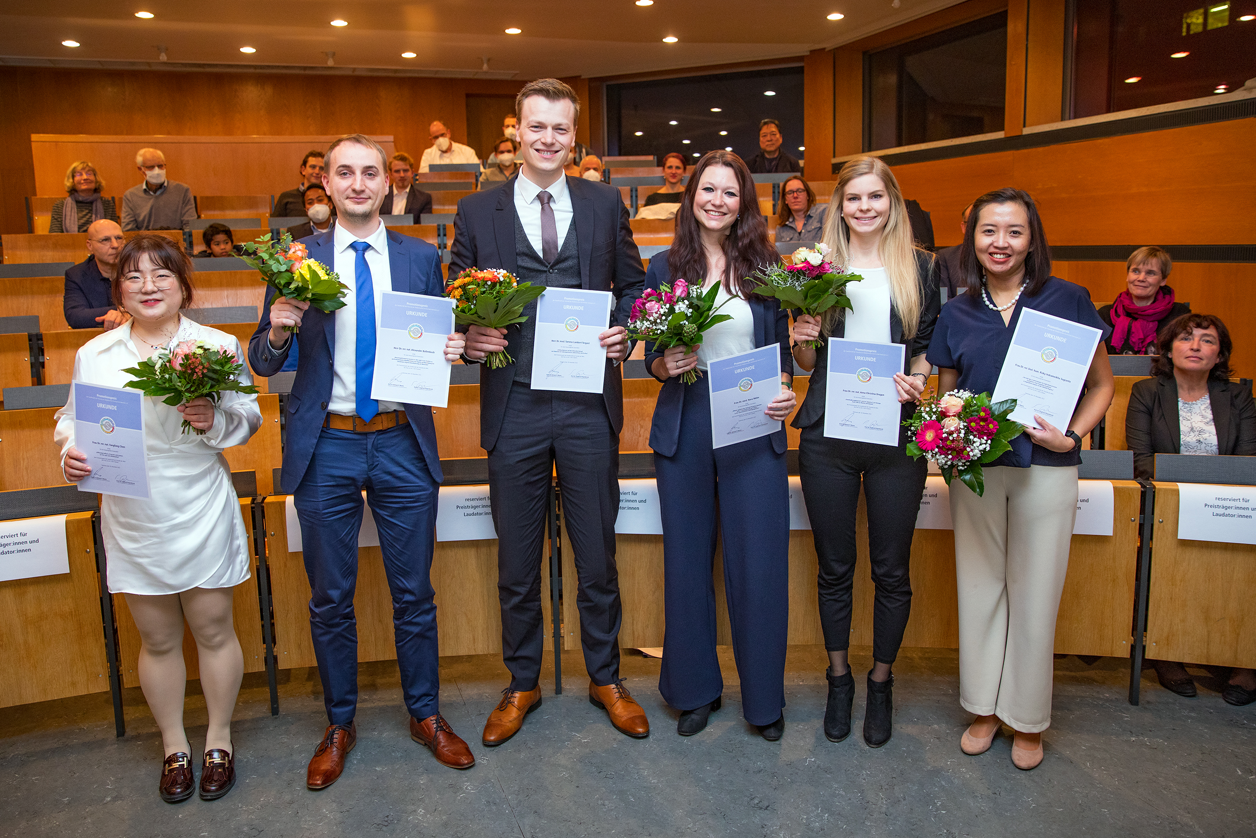 Die Promotionspreisträgerinnen und Promotionspreisträger (von links): Dr. rer. nat. Fangfang Chen, Dr. rer. nat. Alexander Bollenbach, Dr. med. Tammo Lambert Tergast, Dr. med. Nora Möhn, Dr. rer. nat. Anna Christina Dragon und Dr. rer. biol. hum. Rizky Indrameikha Sugianto stehen in einem Hörsaal, hinter ihnen die Sitzbänke.