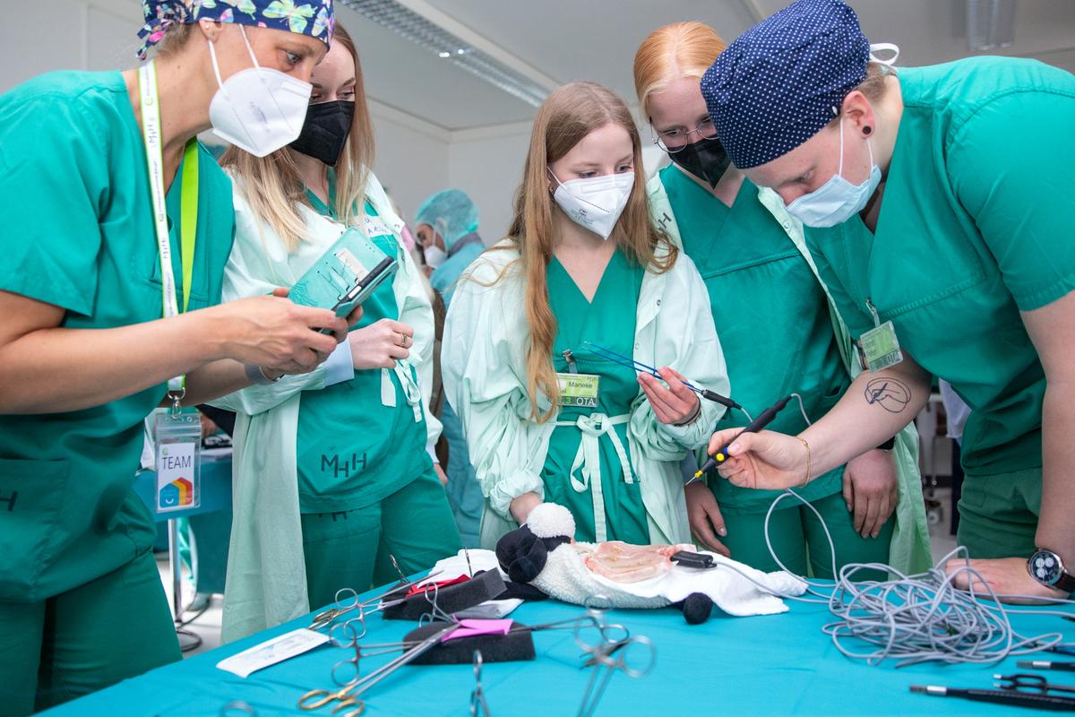 Mehrere junge Menschen in grüner OP-Schutzkleidung stehen um eine Tisch, auf dem ein Schweineschnitzel liegt. Eine Person hält einen Metallstab in der Hand. 