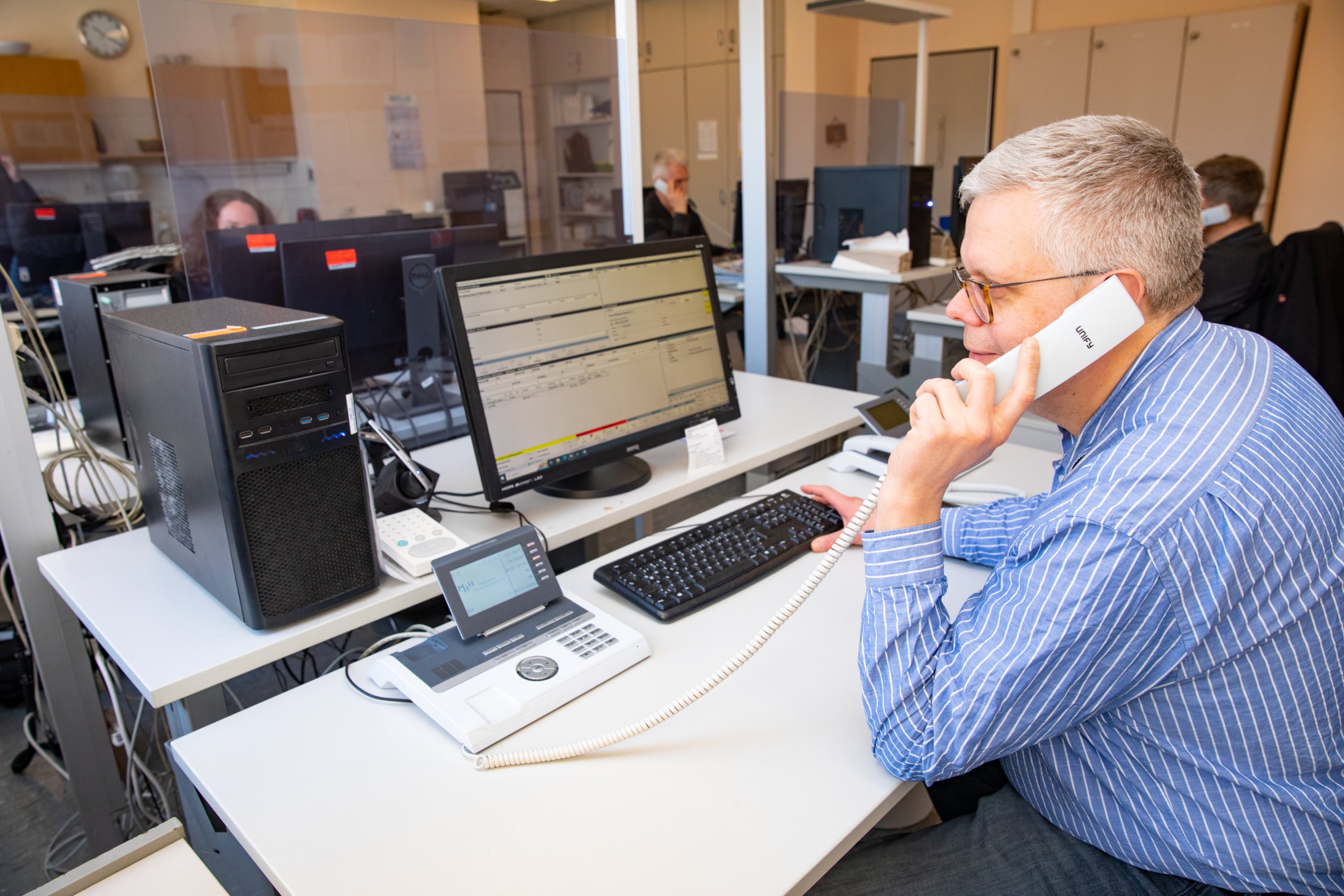 Ein Mann sitzt an einem Schreibtisch mit PC-Monitor und hält einen Telefonhörer am Ohr. 