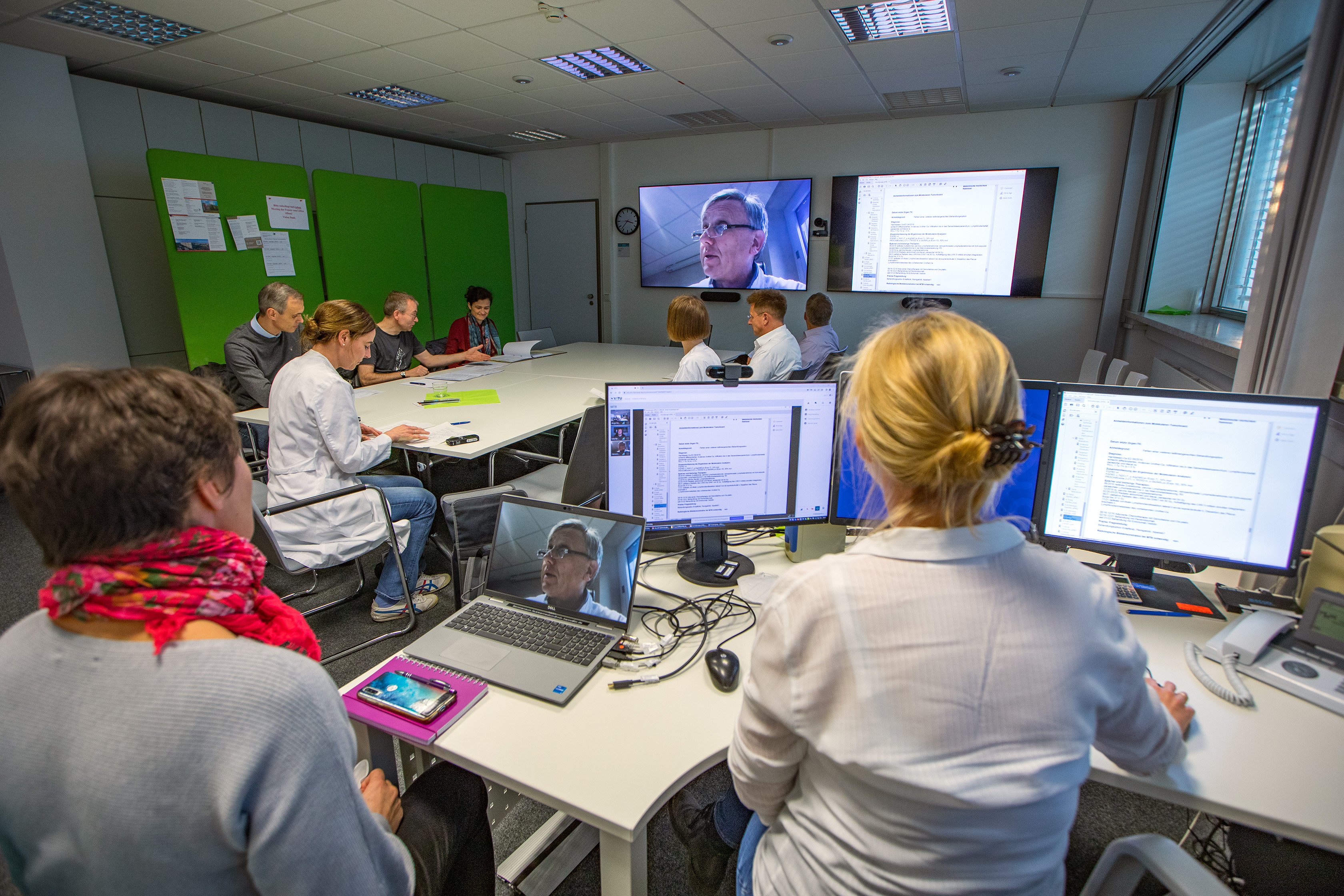 Das Expertenteam, bestehend aus verschiedenen Personen, sitzt in einem Raum, auf Bildschirmen sind Unterlagen zu sehen, eine Person ist über Videokonferenz dazu geschaltet.