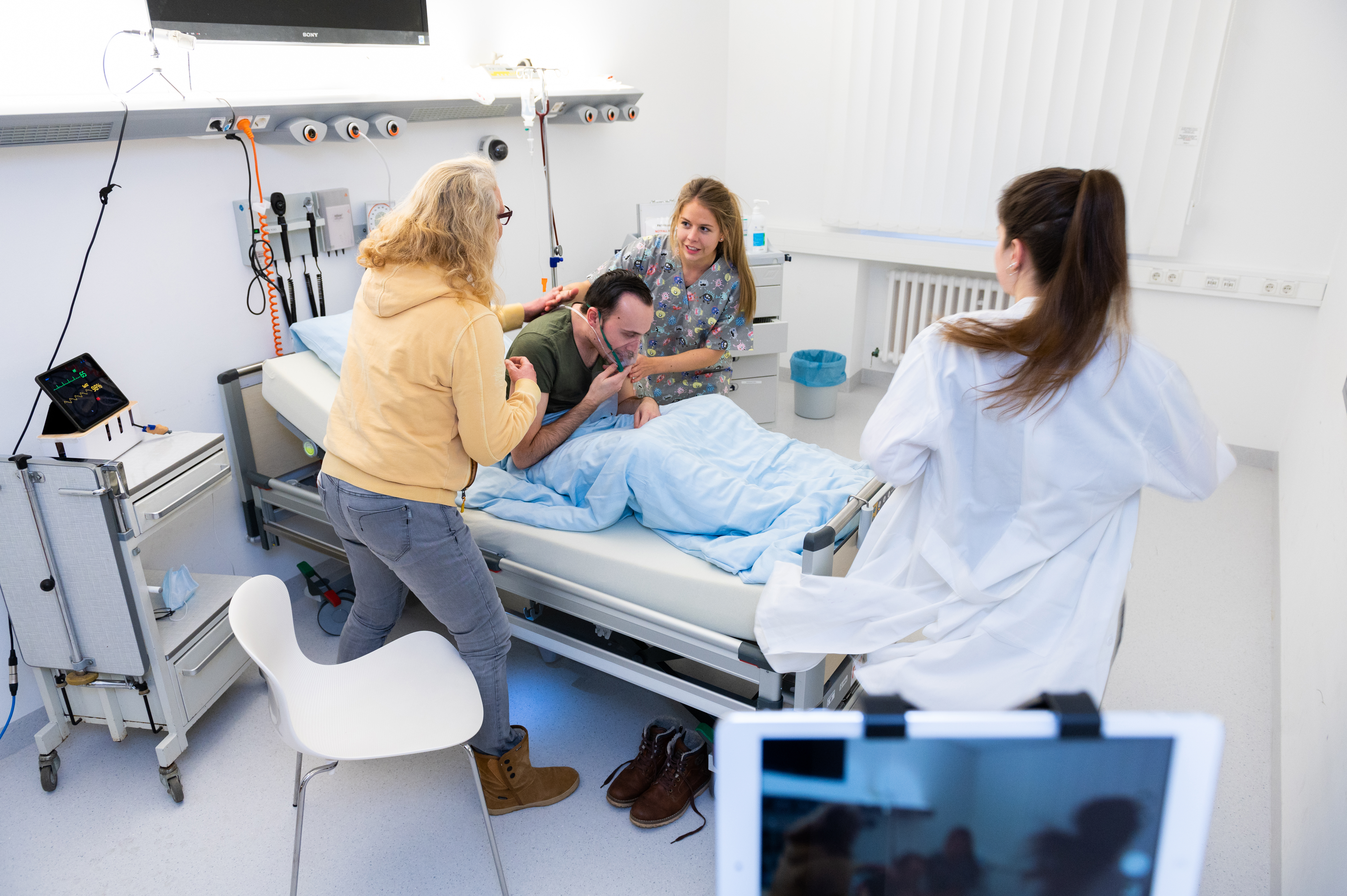Studierende und Pflegeauszubildende stehen um ein Krankenbett, in dem ein Patient mit Atemmaske liegt und üben eine Notfallsituation.