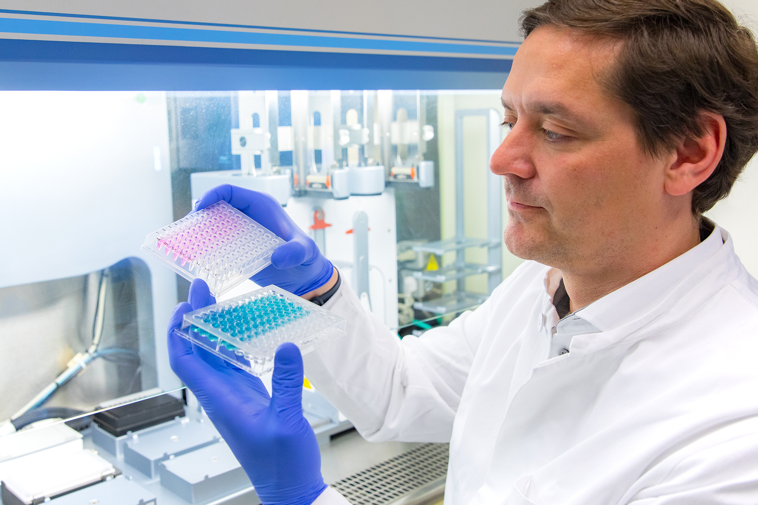 Professor Dr. Christian Bär stands in front of a pipetting robot and holds two micropipetting plates in his hands