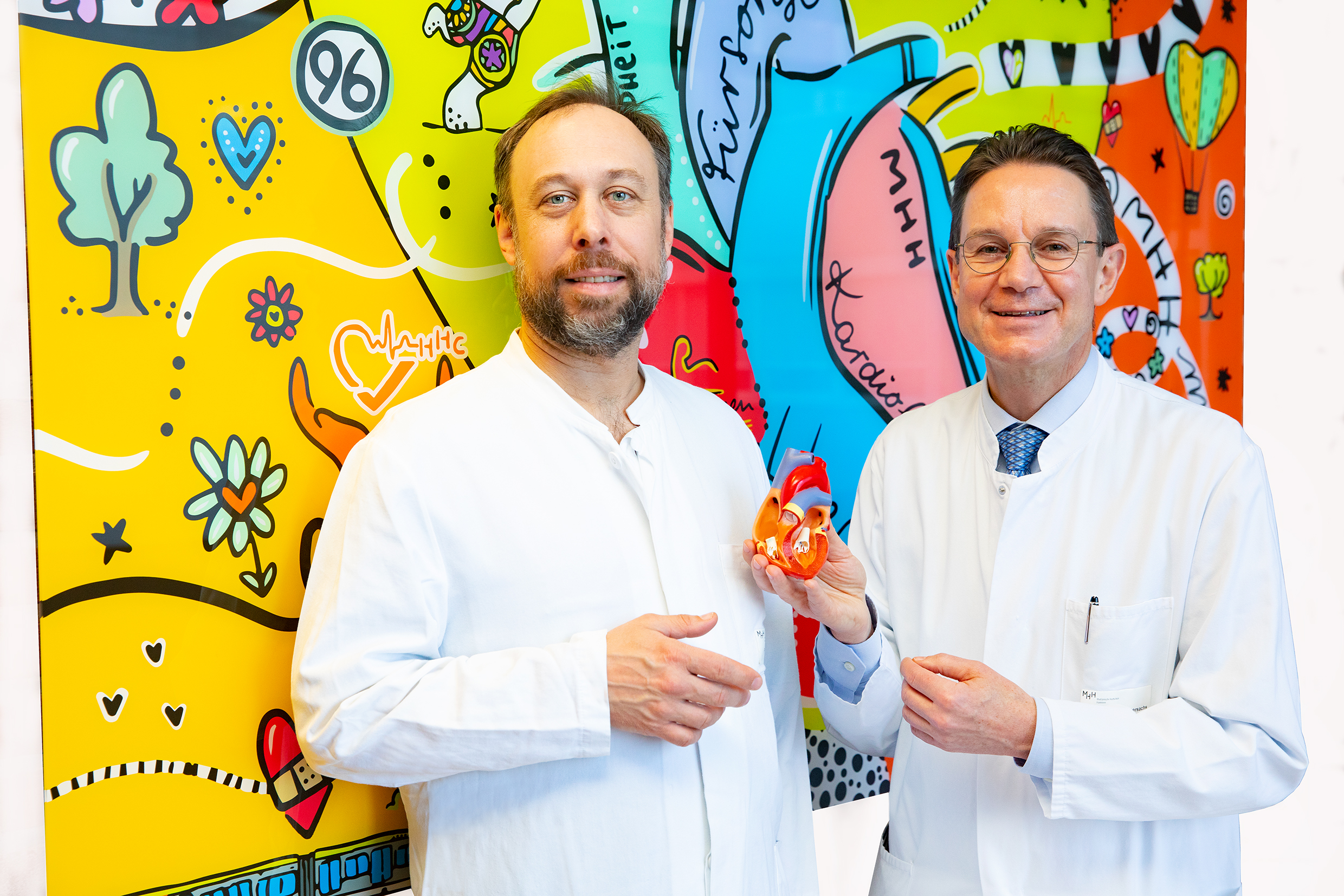 Professor Dr. Tibor Kempf (links) und Professor Dr. Johann Bauersachs stehen vor einer bunt gestalteten Wand, Professor Bauersachs hält ein Herzmodell in der Hand.