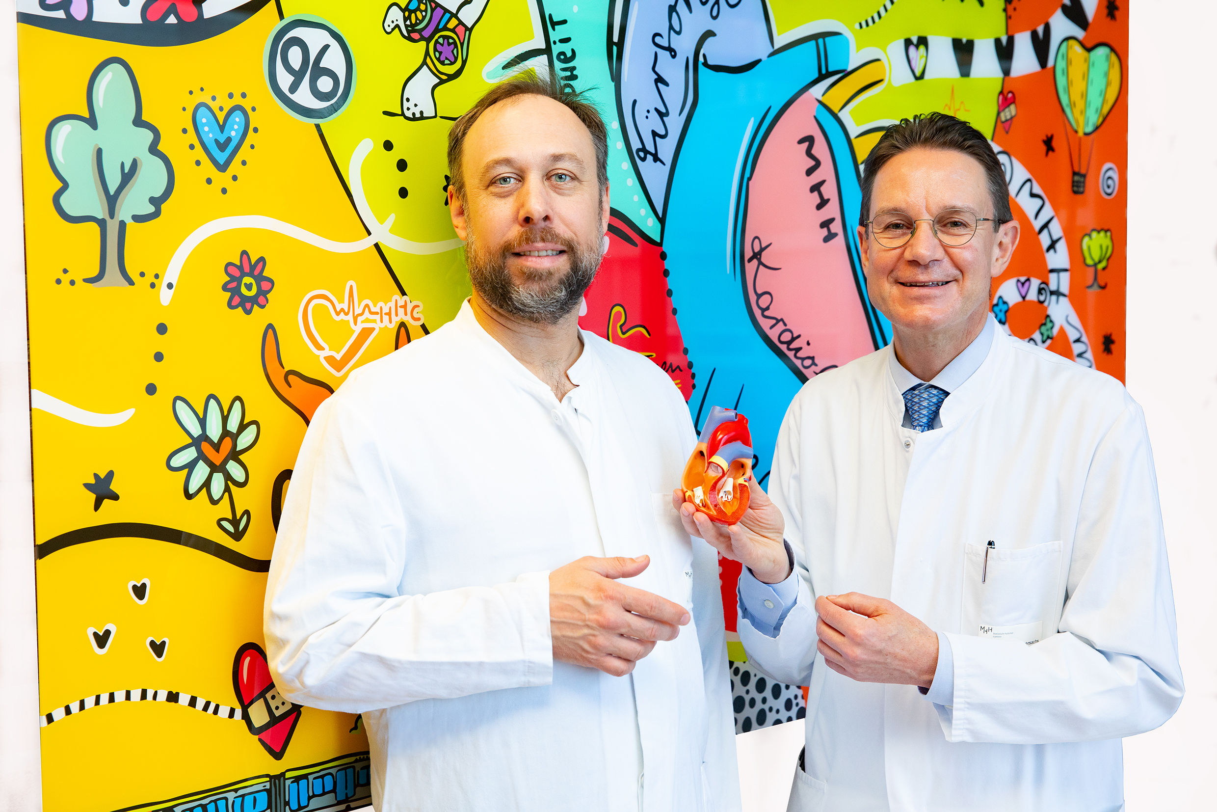 Professor Dr. Tibor Kempf (left) and Professor Dr. Johann Bauersachs stand in front of a colourful wall, while Professor Bauersachs holds a heart model in his hand.