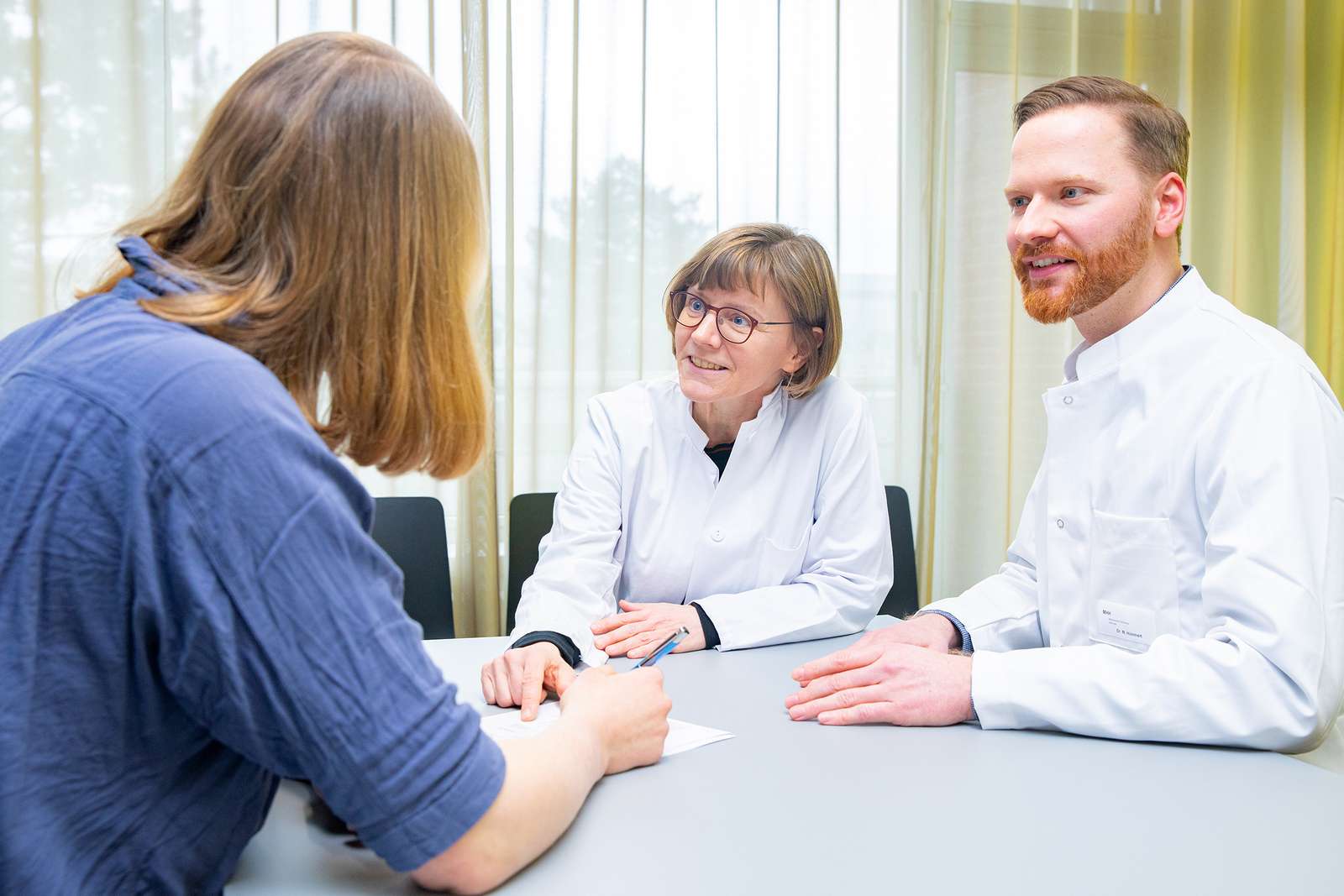 Professorin Trebst und Dr. Hümmert, beide im Arztkittel, sitzen an einem Tisch und unterhalten sich mit einer Patientin, die ihnen gegenübersitzt.