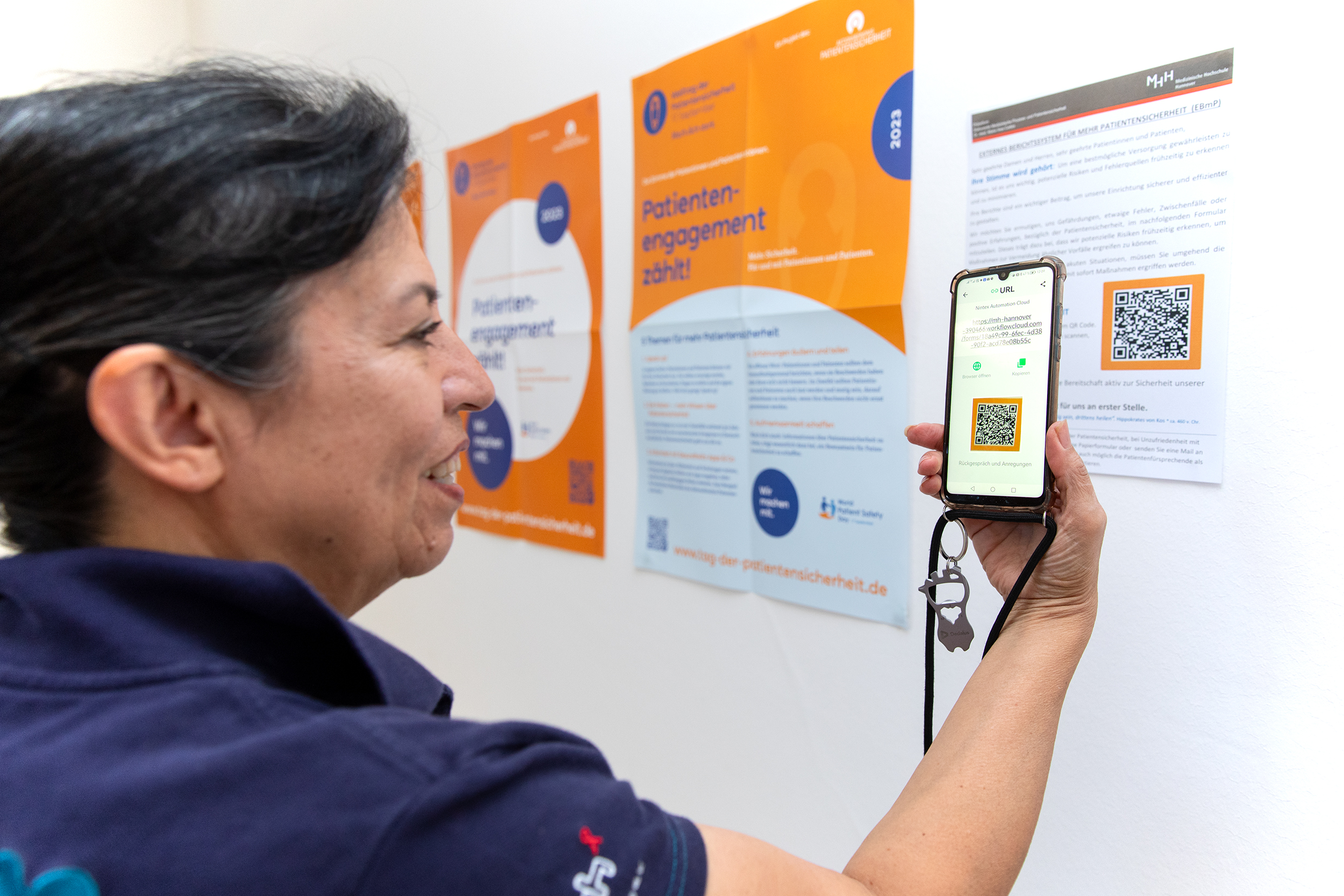 Dr. Maria Cartes stands in front of a poster, which shows the QR Code. She scans it with her smartphone.