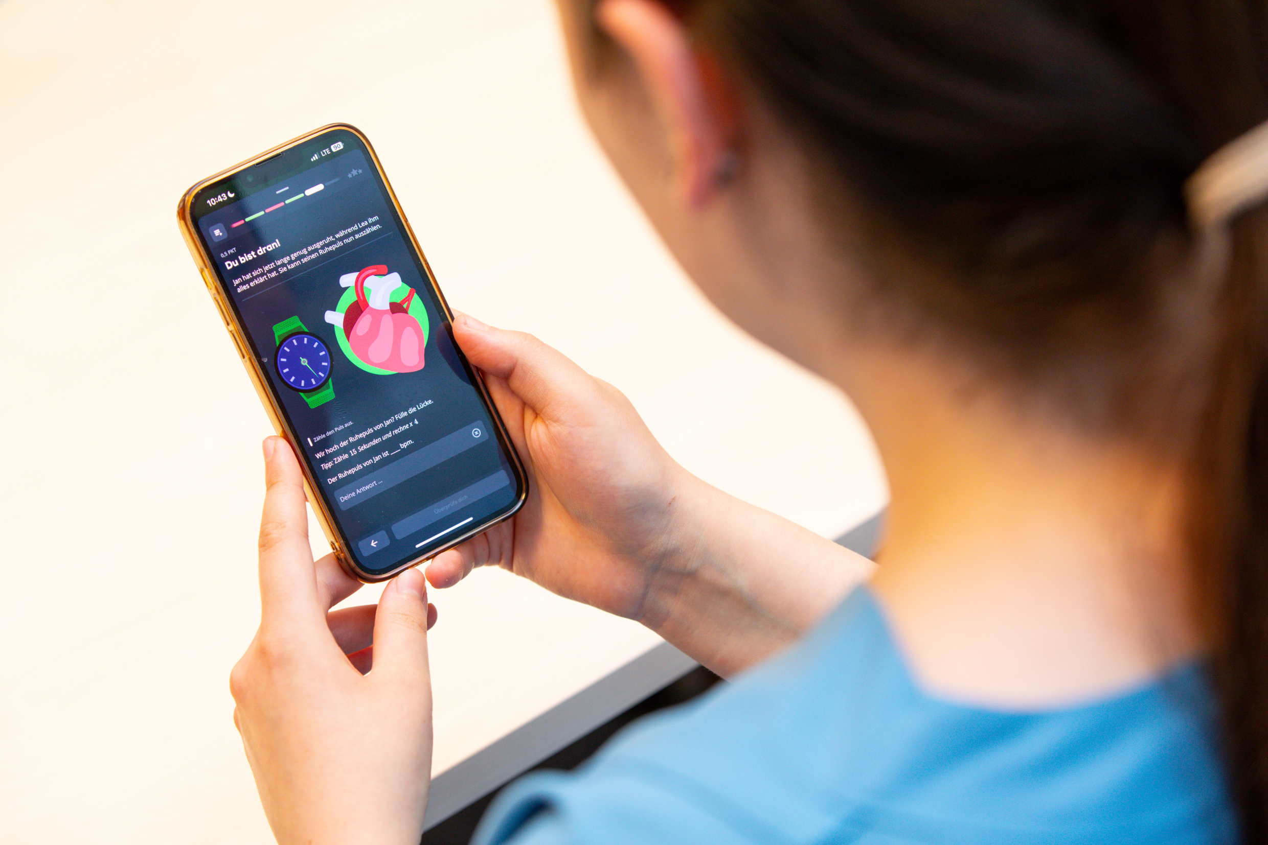 Blick über die Schulter einer jungen Frau mit dunklen Haaren hinunter auf ein Smartphone, das einen Ausschnitt aus der App zeigt.