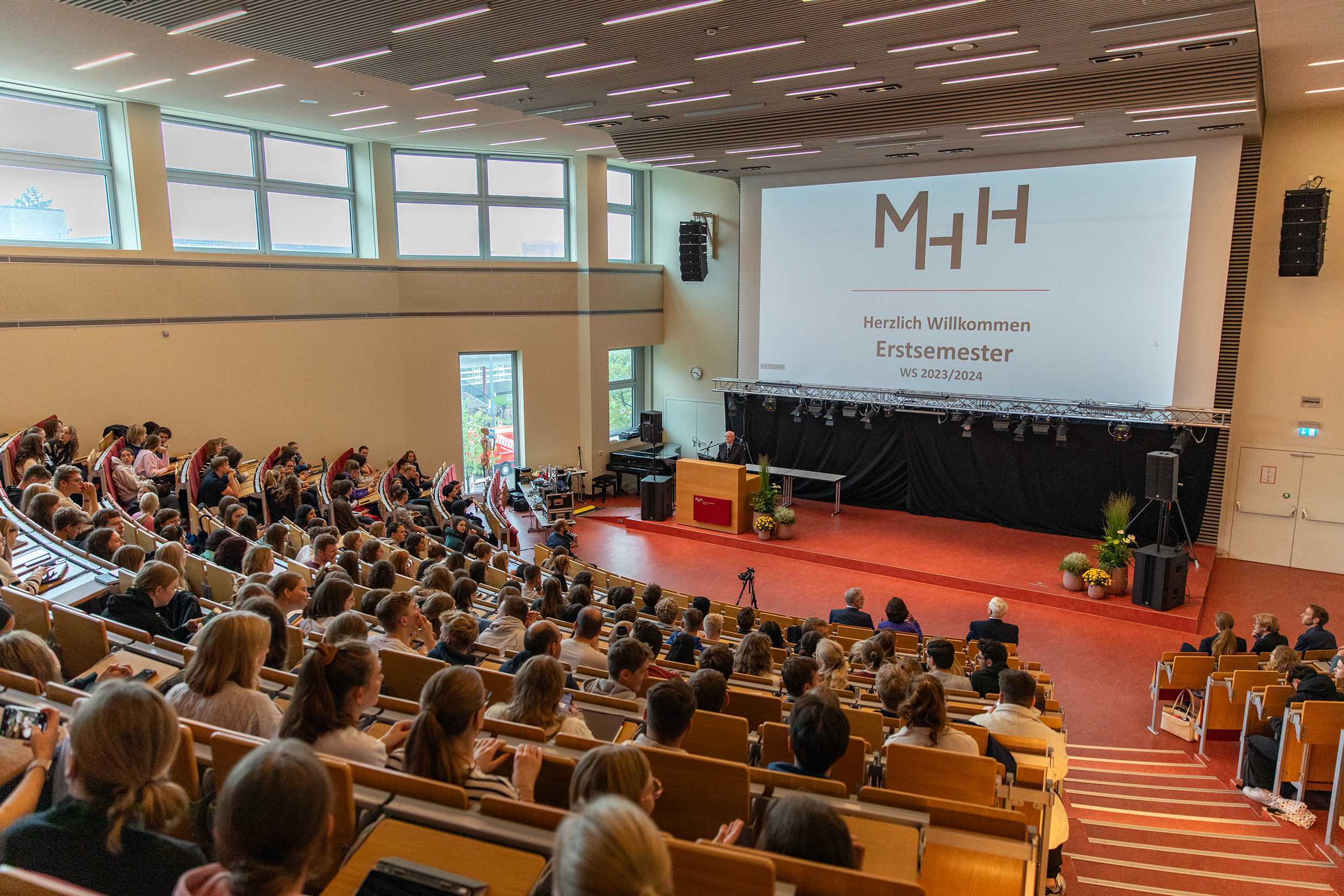 ein Foto der Erstsemester, die in einem Hörsaal sitzen. Fotografiert wurde vom oberen Bereich des Hörsaals nach unten. Ein Beamer projeziert die Worte "Herzlich Willkommen Erstsemester" an die WandWorte