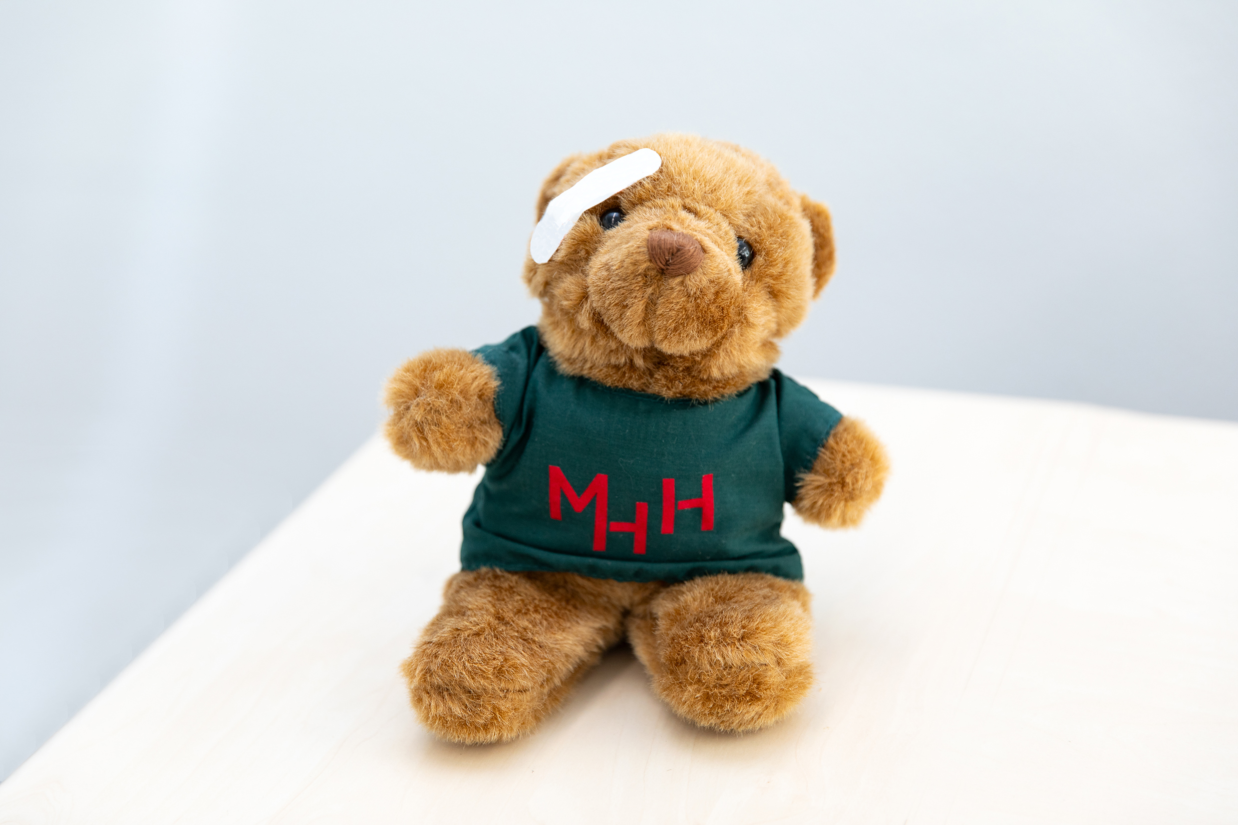 ein Teddybär trägt ein dunkelgrünes T-Shirt mit rotem MHH-Aufdruck. Über seinem Auge klebt ein kleines weißes Pflaster