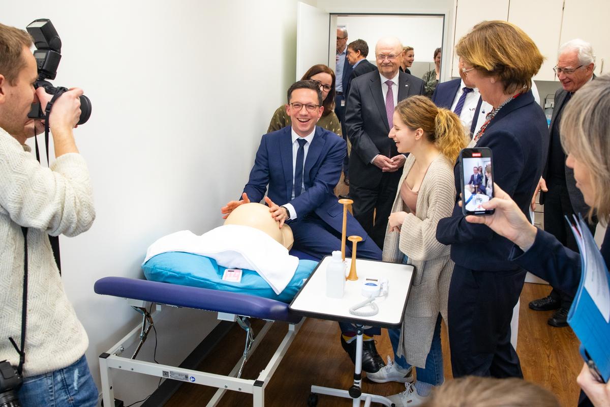 Falko Mohrs sits on a lounger and holds his hands on a dummy stomach. Press representatives, midwives and MHH President Michael Manns stand around him.