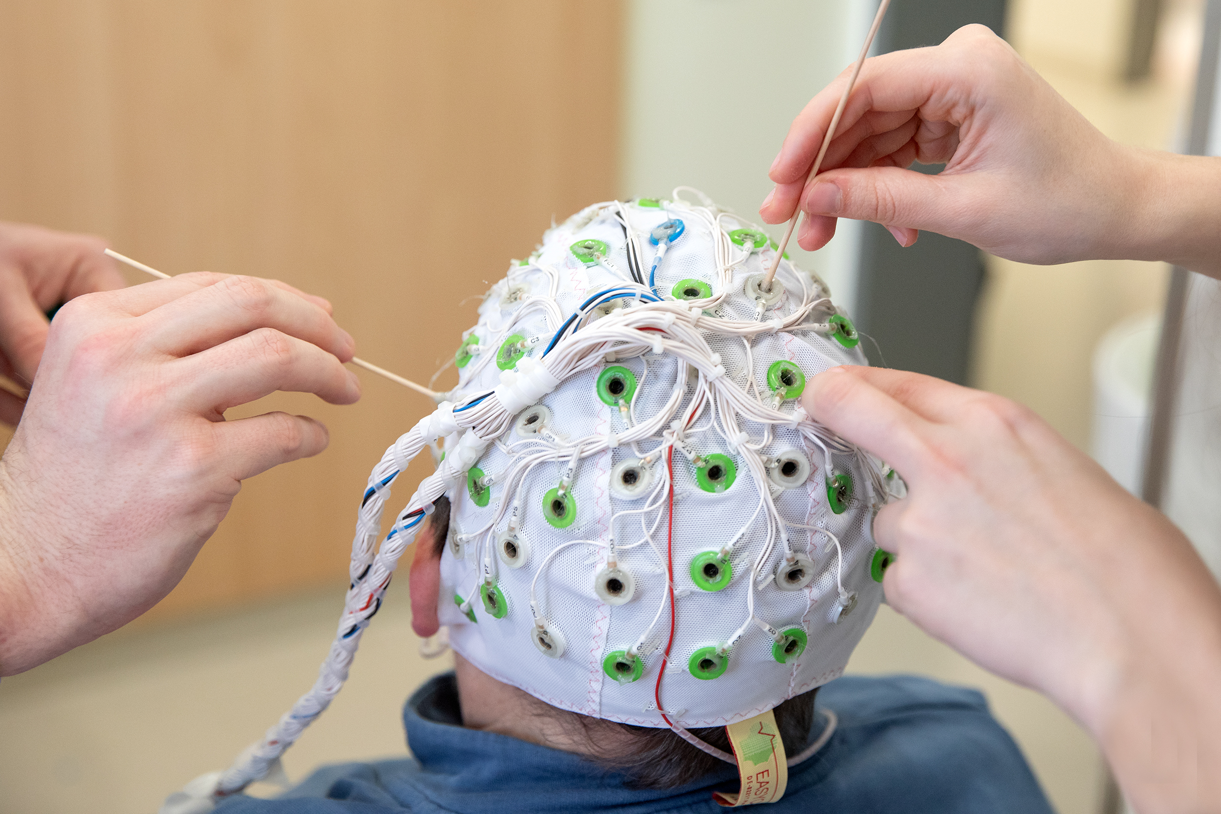 Two pairs of hands apply contact gel to the electrodes of an EEG cap.