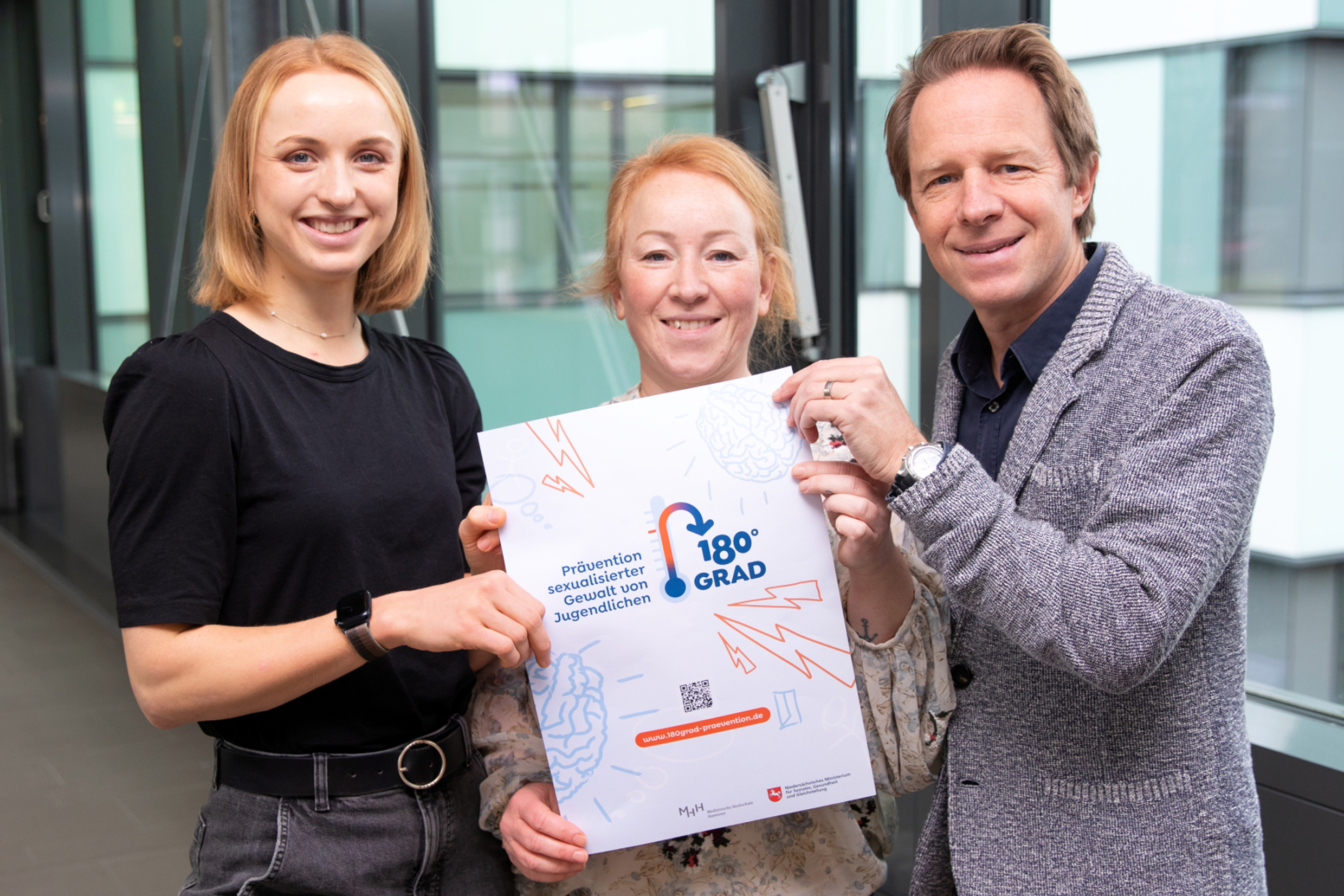 Zwei Frauen und ein Mann stehen mit einem Plakat in der Hand in einem Klinikgang.