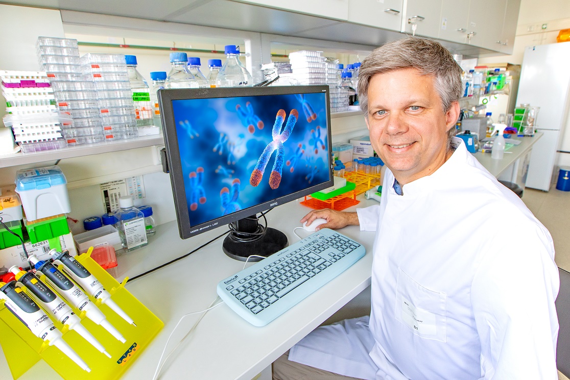 Auf dem Foto ist Prof. Dölken im weißen Laborkittel zu sehen, er sitzt auf einem Stuhl in einem Labor und auf dem Tisch neben ihm steht ein Bildschirm.