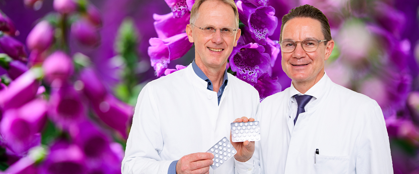 Zwei Männer im weißen Kittel stehen vor einem Foto mit rotem Fingerhut und halten Tabletten-Blister in der Hand.