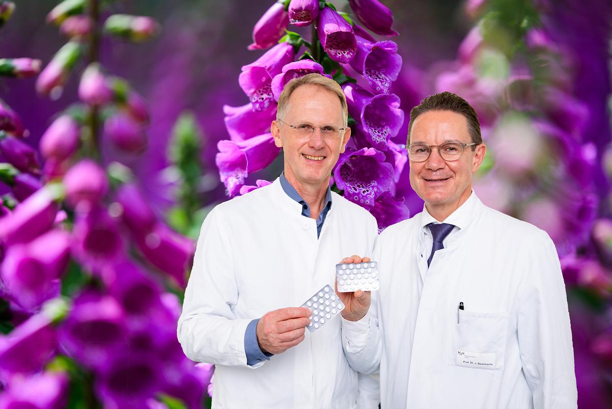 Zwei Männer im weißen Kittel stehen vor einem Foto mit rotem Fingerhut und halten Tabletten-Blister in der Hand.