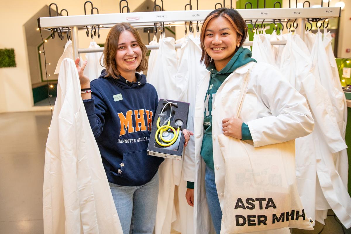 Two women are standing in a cloakroom with lots of white coats hanging on it, presenting the First Semester Student bags and their contents.