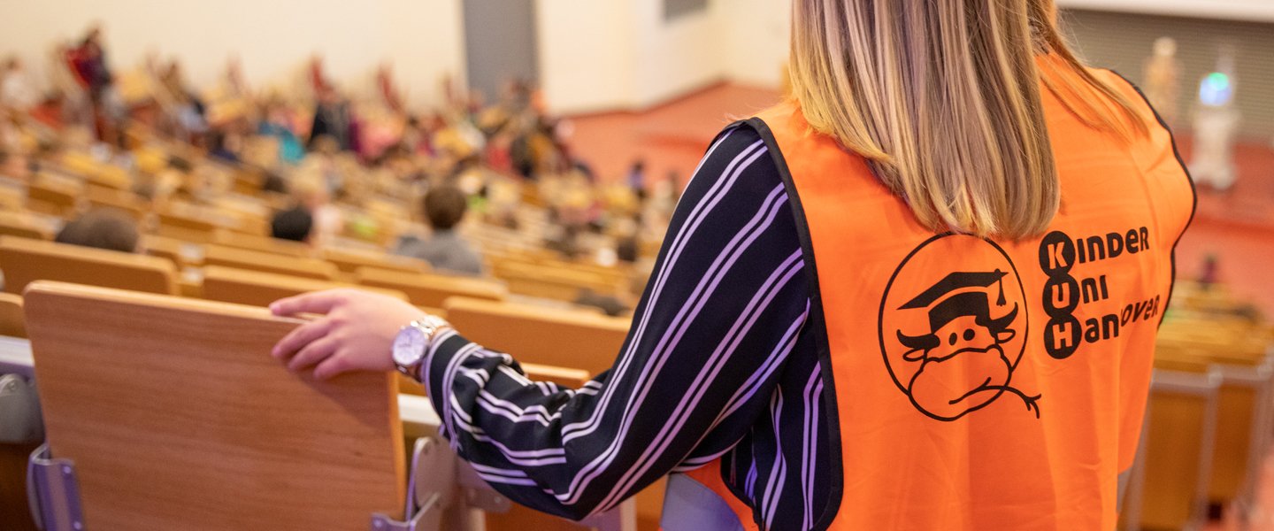 A person with an orange vest looks down from above in a lecture hall. The KinderUni logo is on the vest.