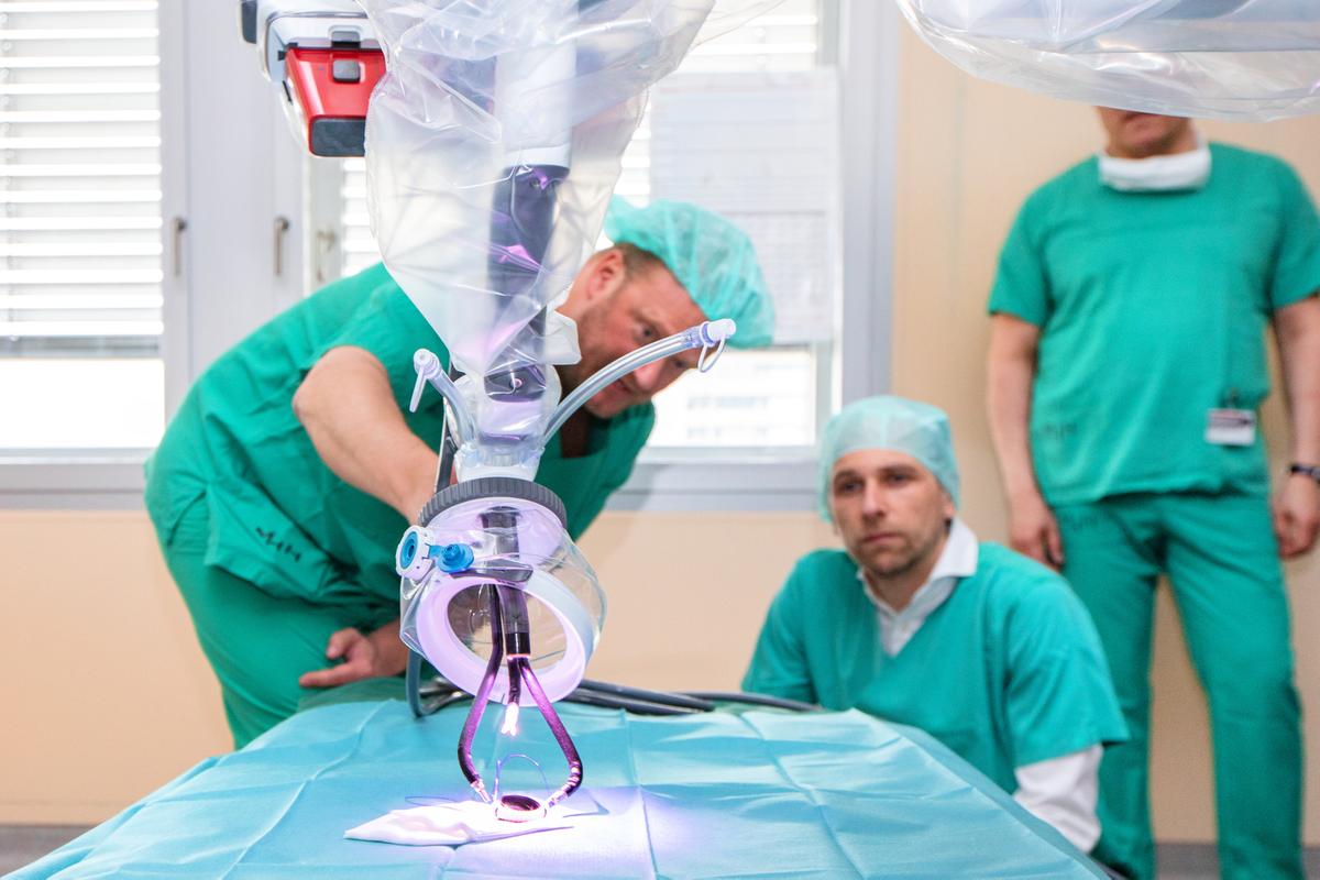 Two men in surgical scrubs are observing the arm of a surgical robot 