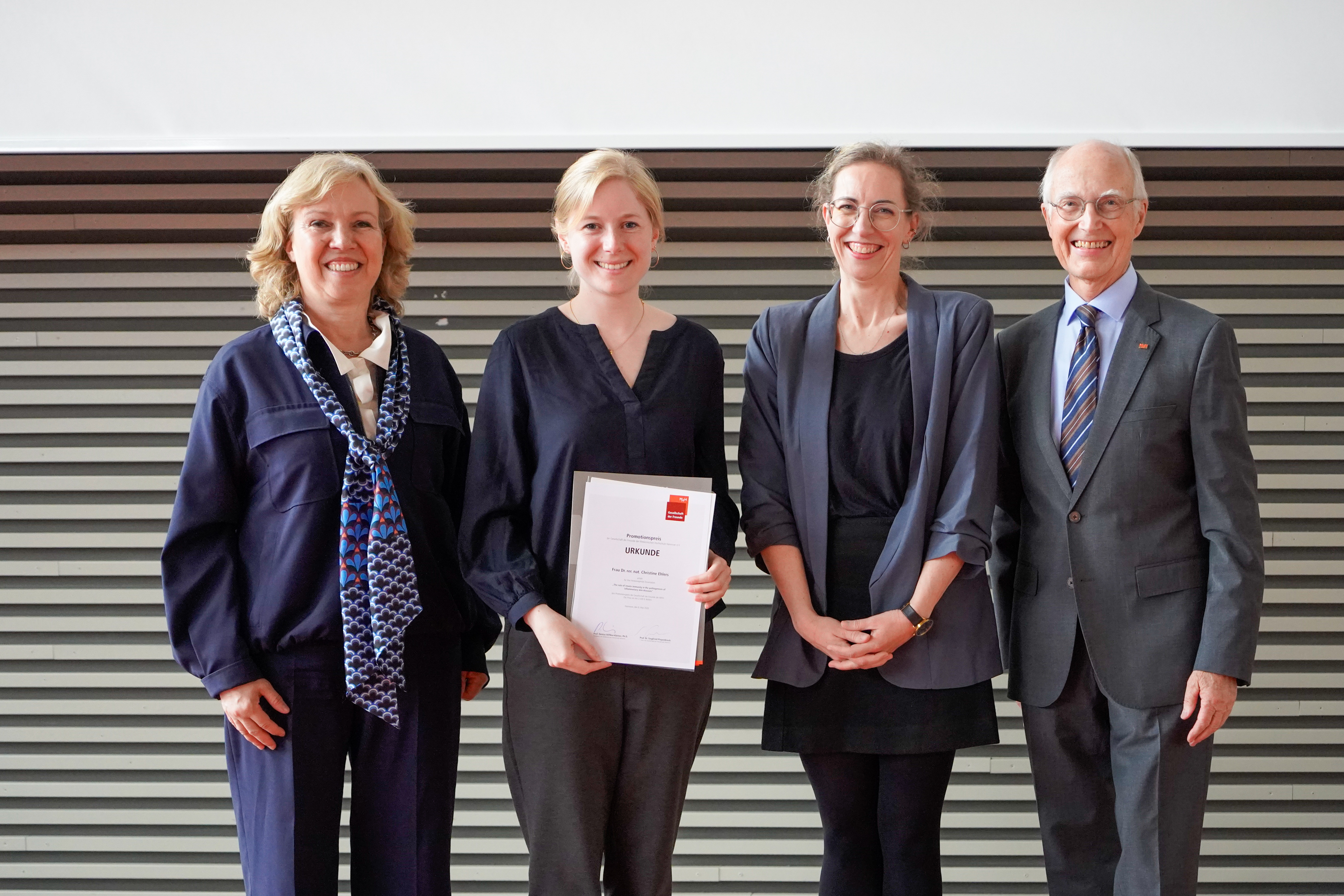 Eine Promotionspreisträgerin präsentiert ihre Urkunde neben drei Hochschulvertreter:innen bei der Promotionsfeier.