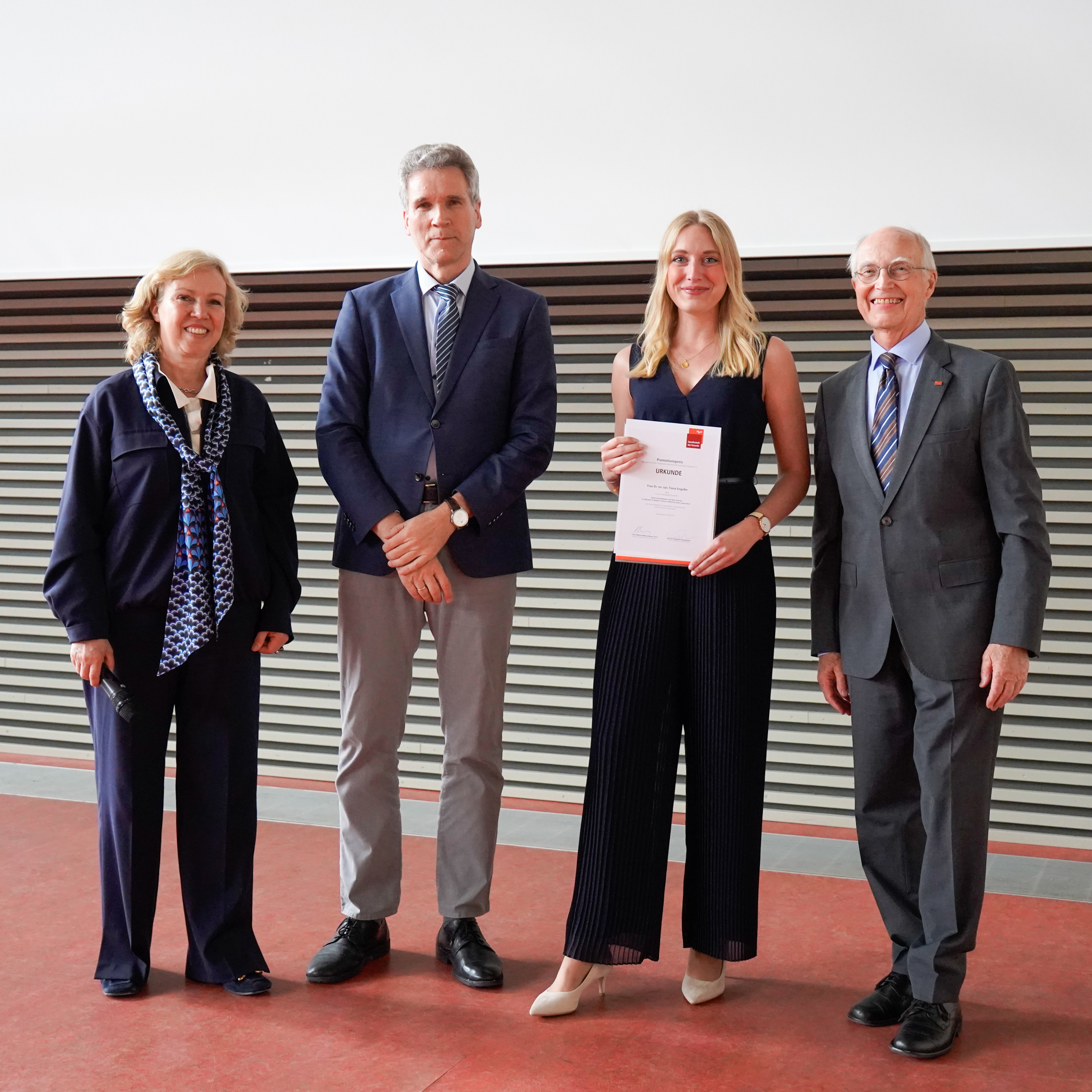 Eine Promotionspreisträgerin präsentiert ihre Urkunde neben drei Hochschulvertreter:innen bei der Promotionsfeier.
