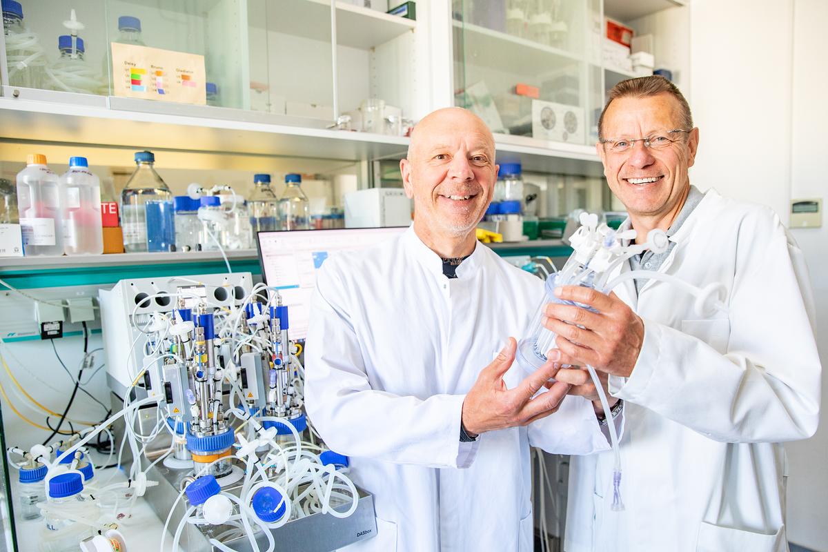 Zwei Wissenschaftler in weißen Kitteln stehen in einem Labor und halten einen Bioreaktor in den Händen.