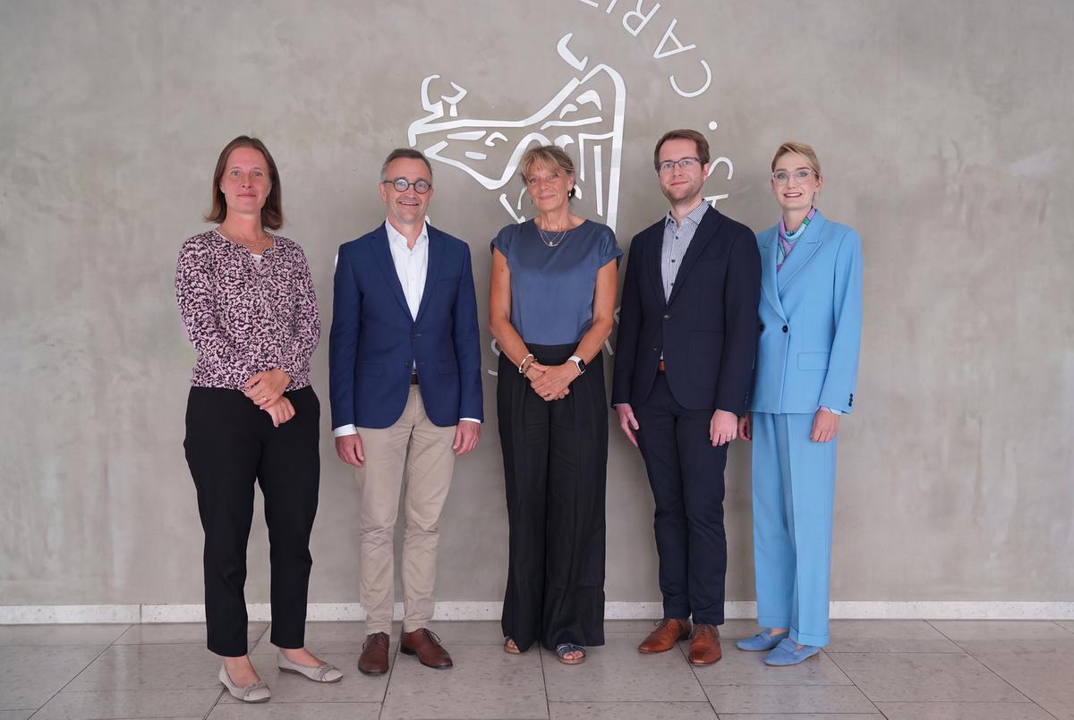 Fünf Personen stehen vor einer Wand mit einem kunstvollen Schriftzug und einem grafischen Motiv. Die Gruppe blickt lächelnd in die Kamera.