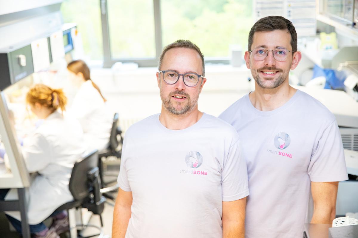 Zwei Männer stehen nebeneinander im Labor und lächeln freundlich in die Kamera. Im Hintergrund sind Personen zu sehen, die an einer Laborbank arbeiten.