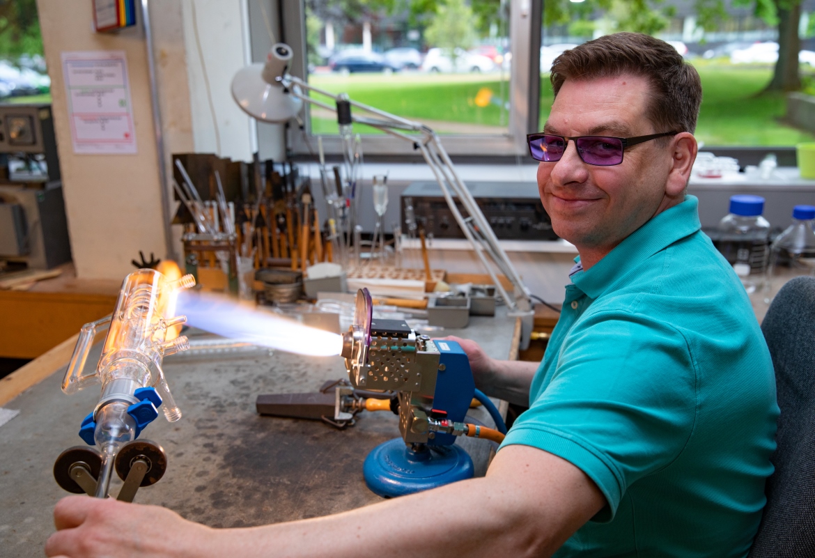 Ein Mann sitzt am Arbeitstisch und bedient einen Brenner, um Glas zu formen. 