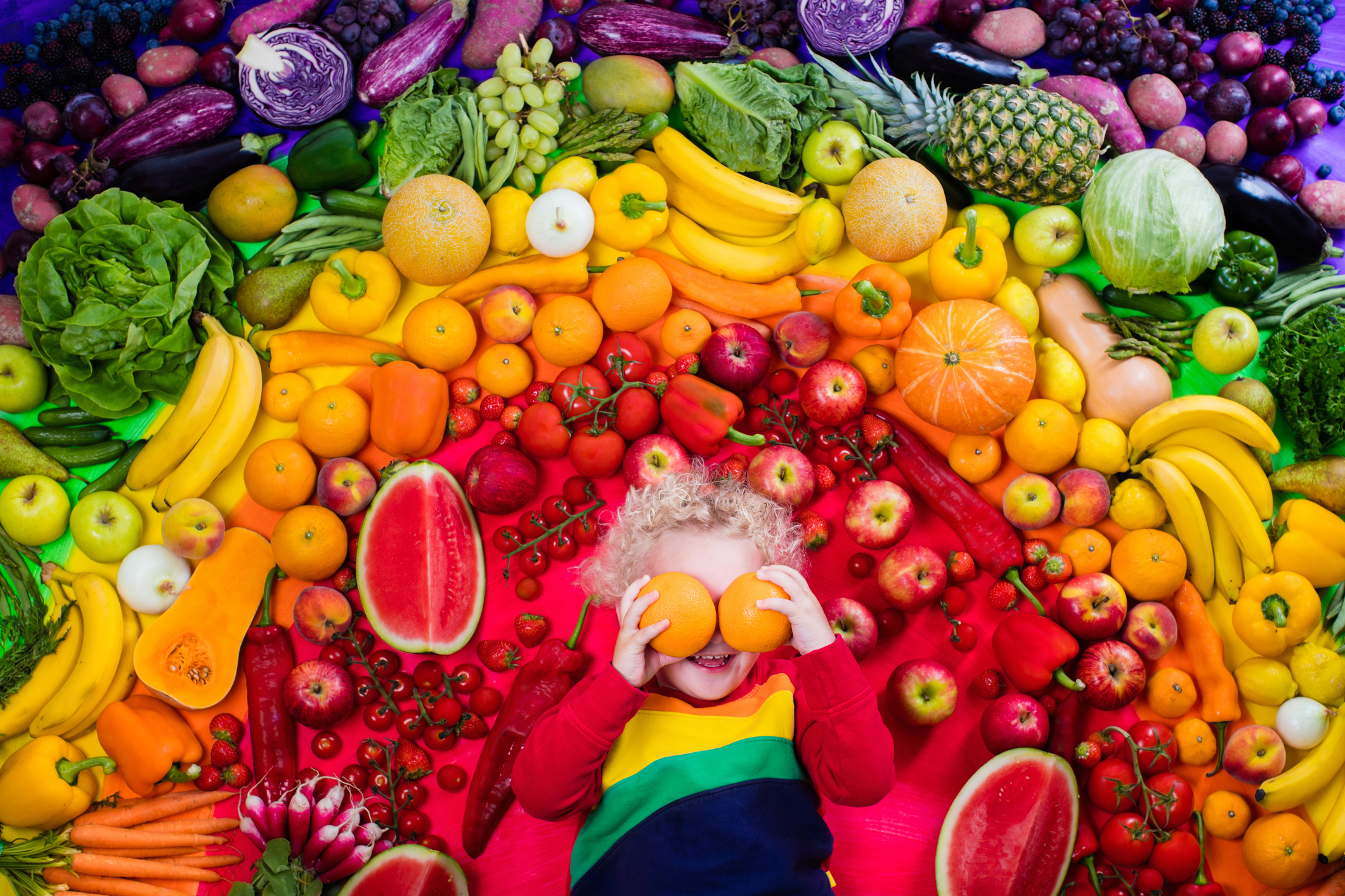 Kleines blondes Mädchen mit Orangen vor den Augen und viel Obst und Gemüse in Regenbogenfarben 