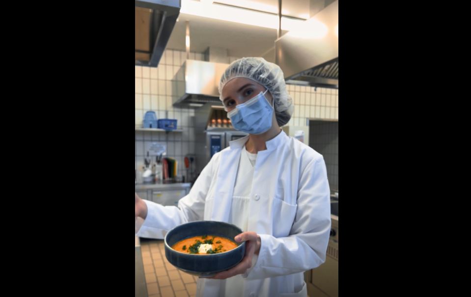 Eine junge Frau mit weißem Schutzkittel und Maske hält einen Teller mit Möhrensuppe in der Hand. 