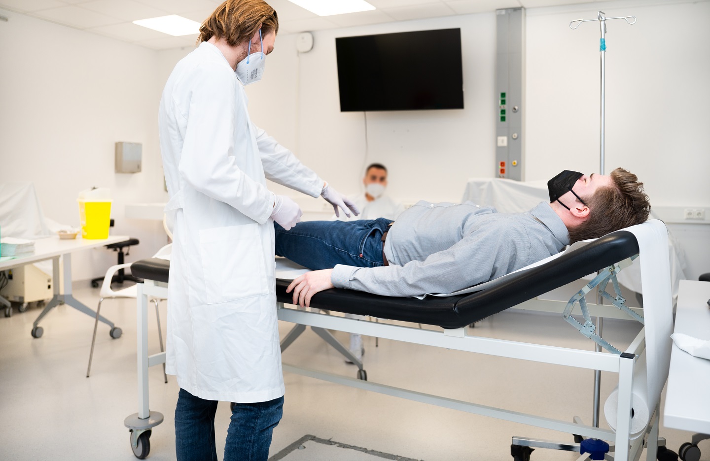 Studierende untersucht bei einer Prüfung einen Simulationspatienten auf einer Liege. Prüfer sitzt im Hintergrund.