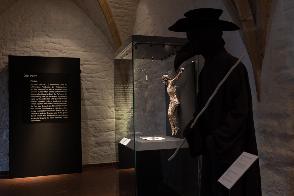 Im Vordergrund ist das typische Gewand der sogenannten Pestdoktoren zu sehen: großer Hut, eine Maske mit einem langen Schnabel und ein langer dunkler Umhang.