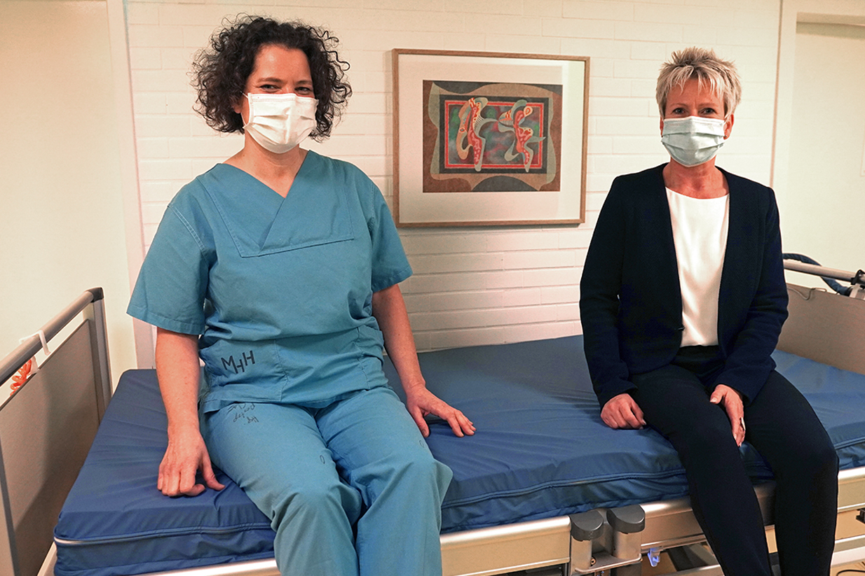 Beeke Luise Schmeling und Dr. Regina Schmeer sitzen mit Mund-Nasen-Bedeckung auf der neuen Matratze.