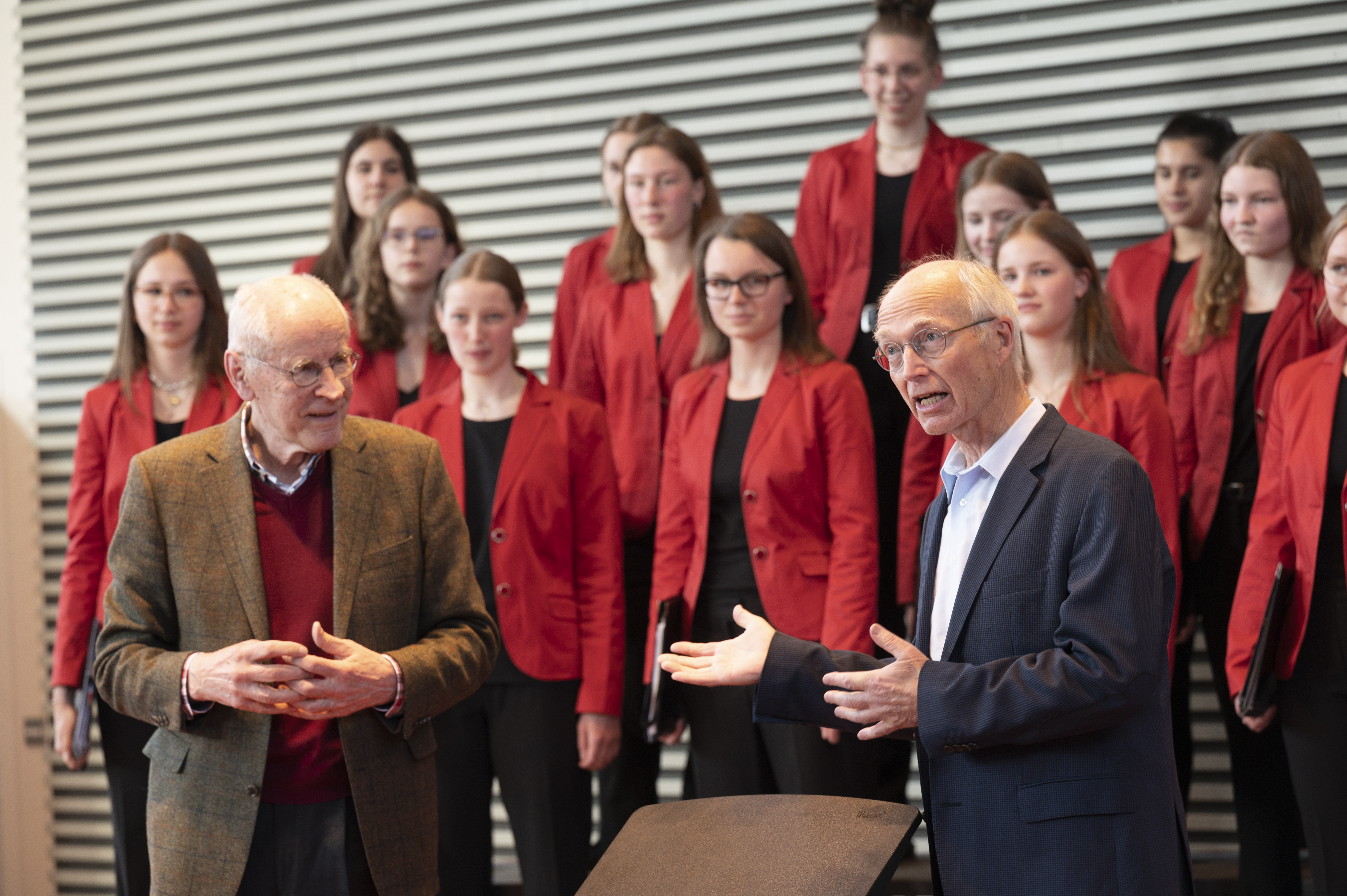 Die Professoren stehen gemeinsam im Vordergrund, hinten ist undeutlich der Mädchenchor zu erkennen.