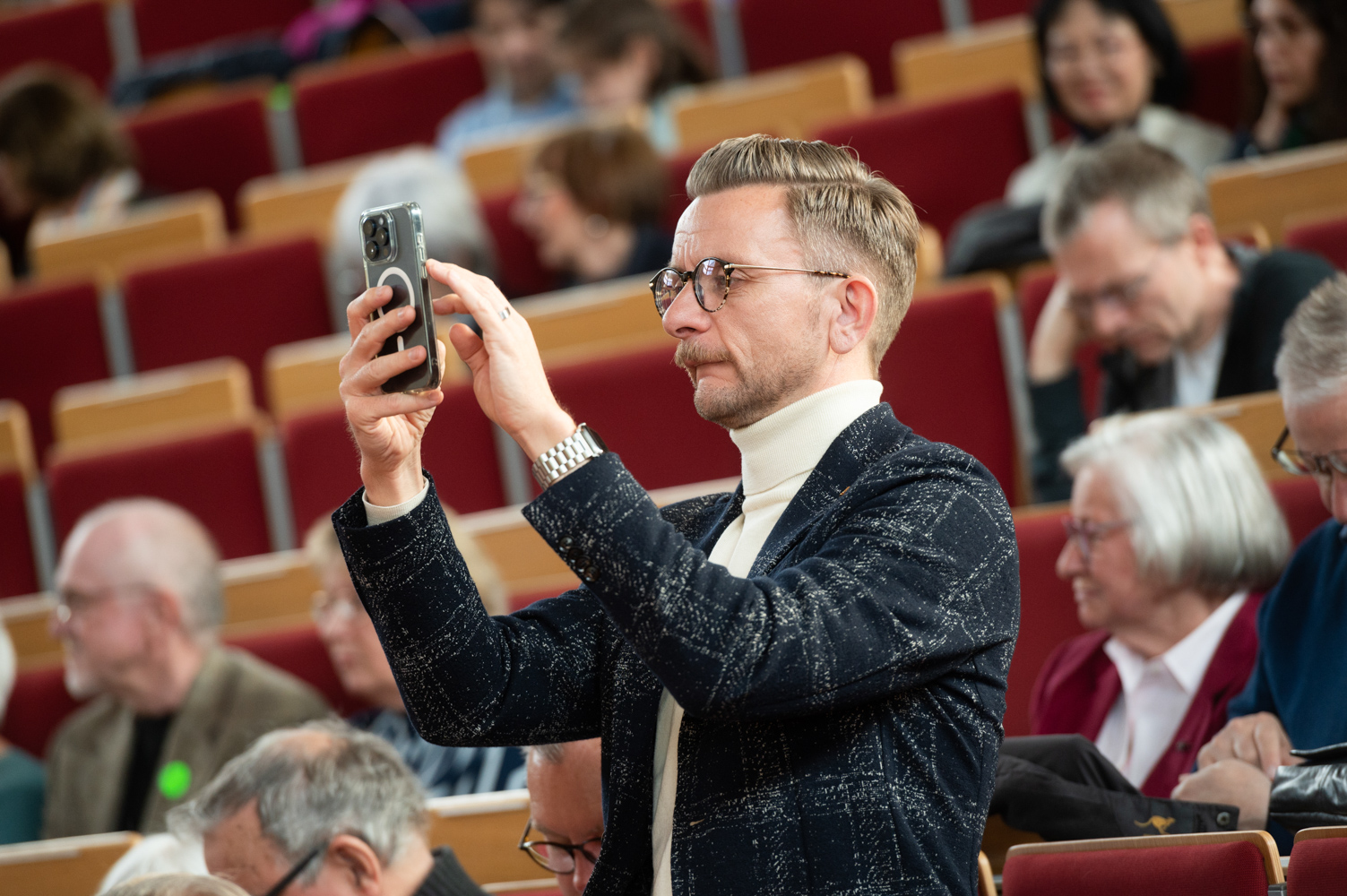 Ein Gast steht in den Stuhlreihen von Hörsaal F und macht mit seinem Handy ein Foto.