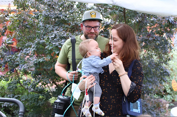 Theos Familie im Garten. Theo bekommt über einen Schlauch Sauerstoff. Sowohl Mutter als auch Vater tragen Geräte, damit theo Sauerstoff bekommen kann.  Copyright: privat / Theos Familie