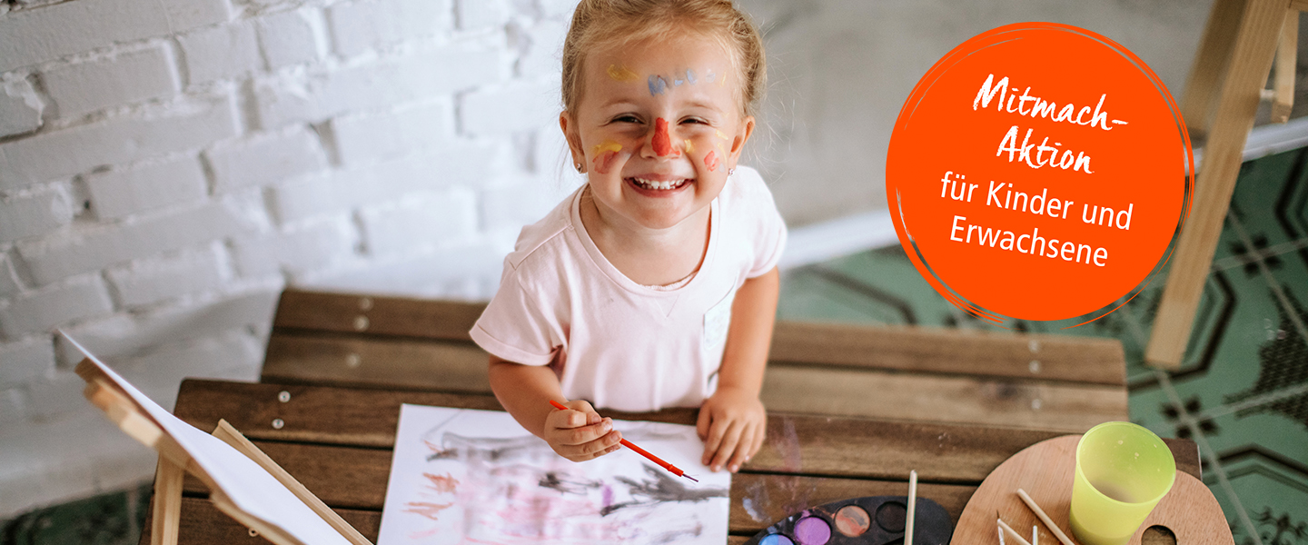 Ein junges Mädchen mit roten Tupfer auf der Nase und Pinsel in der Hand malt an einem Tisch und freut sich dabei. Ein roter Button wirbt für die Mitmach-Aktion für Kinder und Erwachsene; Copyright: iStock/StefaNikolic