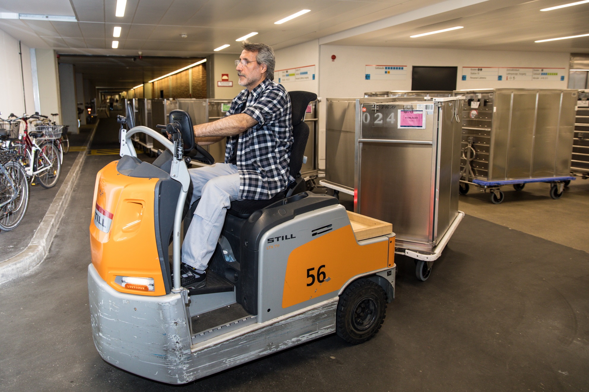 Mitarbeiter des Transport auf einer Elektrolok mit mehreren angekoppelten Transportwagen fährt auf einem Flur der MHH um die Kurve und im Hintergund stehen weitere Transportwagen zur Abholung bereit