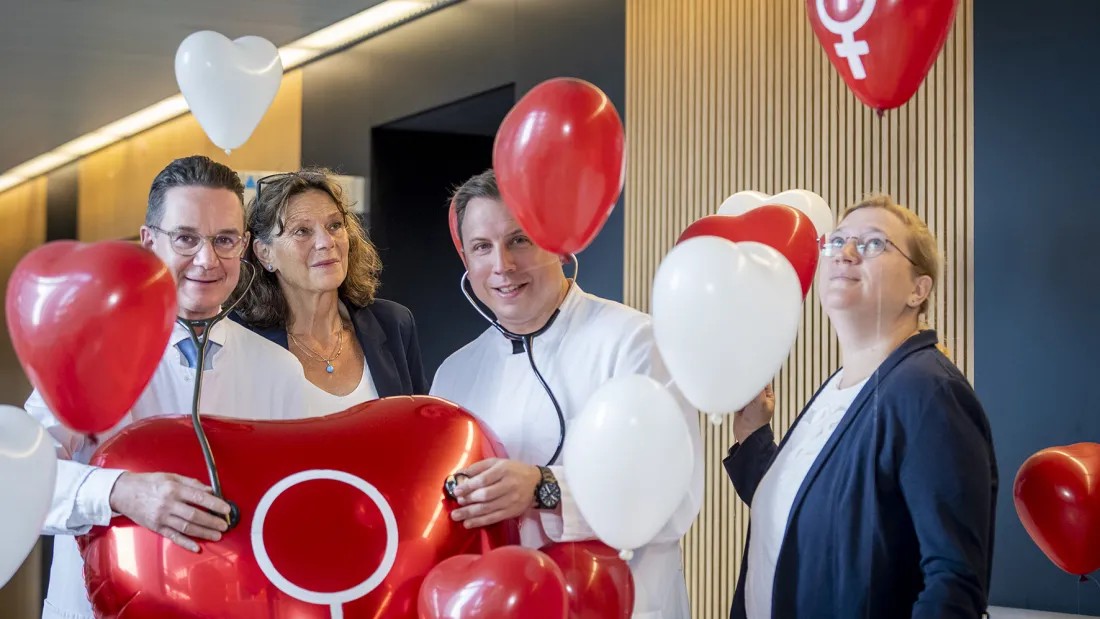 vier Personen, zwei in Arztkittel mit Stethoskop stehen umringt von kleinen Herzballons an einem großen Herzballon