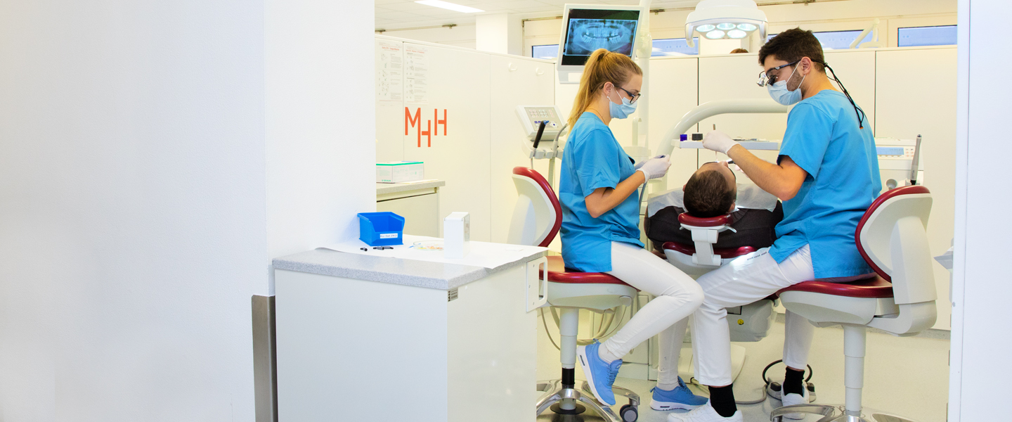 Zwei Studenten (männlich und weiblich) sitzen in blau-weißer Klinikkleidung in einer Behandlungsbox. Der junge Mann schaut in den Mund seines Patienten, der auf dem Behandlungsstuhl liegt, während die assistierende Studentin Instrumentarium vorbereitet 