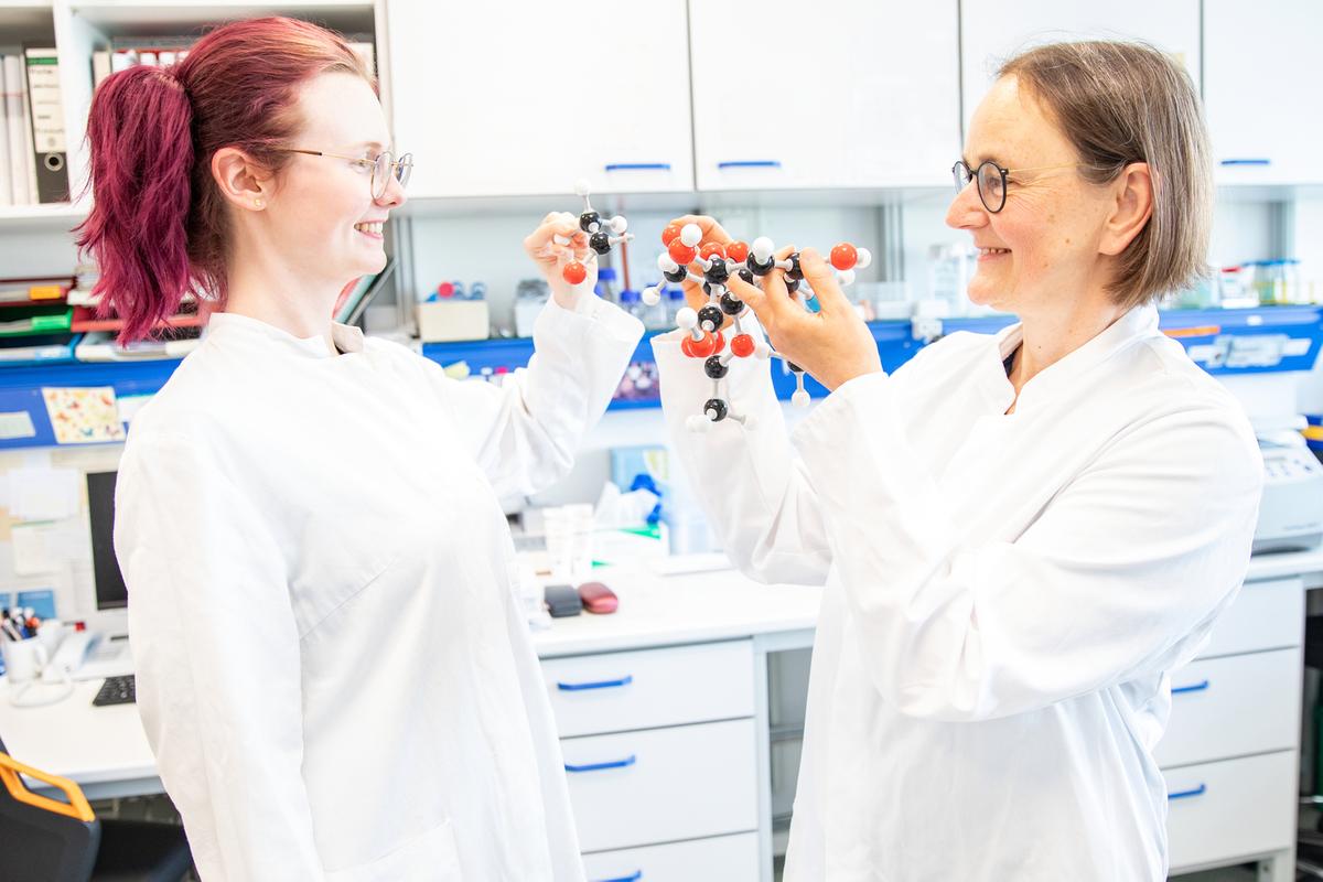 Two female scientists are standing in a laboratory, holding molecular models of a sugar molecule in their hands.